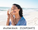 Beautiful latin woman hearing the sound of the sea with a big seashell at beach. Hispanic girl on seacoast with a cockleshell in hands with copy space. Woman on a white sand beach holding a sea shell.