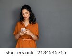 Small photo of Happy young multiethnic woman writing message on smartphone isolated against grey wall. Beautiful smiling hispanic woman checking mail on mobile phone. Cheerful latin girl chatting by cellphone.