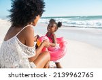 Rear view of young african american woman holding hands of daughter playing with flamingo lifebuoy at beach. Lovely bigger sister dancing with cute little female kid wearing inflatable toy.