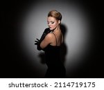 Small photo of A femme fatale in a black dress with an open back and long velvet gloves. Attractive young blonde with makeup for a celebration, portrait in a spot of light