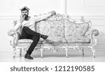 Small photo of Macho sits with open book on head, like roof. Overstudy concept. Man with beard and mustache sits on sofa, white wall background. Guy overdid with studying, fed up of reading old boring book.