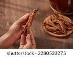 Small photo of A red ginseng root in female hands on a matte brown wooden background. Red ginseng roots are placed on a plate next to the pot of decoction.