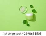 A smear of lotion on a glass platform, a jar of cosmetics and fresh pennywort displayed on a green background. Blank space for branding and advertising.