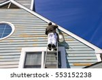 A contractor or painter on a ladder doing exterior paint work, sanding, trim work and repairs on a house, condo or building with wood siding. Blue sky background. Urban city environment. 