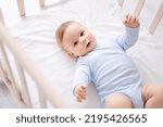 a cute healthy little baby is lying on his back in a crib on white bedding at home in a blue bodysuit. The happy kid looks into the camera, smiles