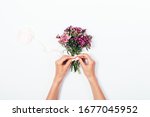 Woman's hands tying satin ribbon bow on small bouquet on white desk, top view.