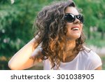 Portrait of a young smiling happy woman in sunglasses at the park on a background of green trees. Girl with curly hair outdoors in the summer.
