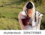 Small photo of Jew wearing tallit and Tefillin or Phylactery praying in nature while covering his face with his hands.