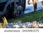 Small photo of Road repaving project, man walking next to an asphalt paving machine with a spray bottle of a very light spray application of diluted asphalt emulsion for a tack coat before laying asphalt
