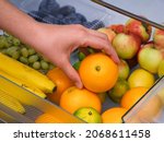 Woman taking orange from fridge drawer full of fruits. Close up. 