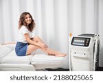 Cheerful young woman smiling at camera, sitting on daybed near diode laser epilation machine. Beautiful woman resting near hair removal device in cosmetology clinic.