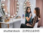 Small photo of Portrait of makeup artist smiling to camera while doing professional makeup in visage studio. Woman sitting at dressing table while female beauty specialist applying lipstick with cosmetic brush.