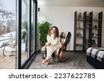 Curly young woman resting inside contemporary barn house in the mountains. Happy female tourist sitting on chair, holding cup of tea, enjoying view of mountain landscape through panoramic windows..