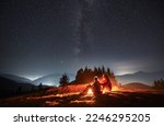 Small photo of Side view of pair sitting one behind other on meadow near bright flame of bonfire and watching surrounding beauty of evening starry sky. Mountain hills and lighted hamlets in the distance.