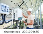 Small photo of Professional worker checking voltage on solar inverter. Male using current probe to measure output voltage. Man in helmet pointing at inveter dial on back side of PV panel.