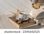 Small photo of Hot coffee cup, book, fall light brown leaves, knitted sweaters in neutral colors on a beige table and white wall background with sunlight shadow. Aesthetic pastel autumn still life.