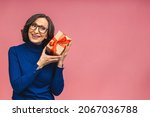 Attractive mature aged aged woman holding presents gift box feels happy studio isolated over pink background. Life events celebration congratulation love care concept