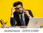 Small photo of Tired sleepy indian businessman talking on mobile phone with client, making online conversation at office workplace desk. Disinterested freelancer hindu man having annoyed boring talk on smartphone