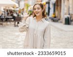 Small photo of Hello. Young pretty woman smiling friendly at camera, waving hands gesturing hi, greeting or goodbye, welcoming with hospitable expression outdoors. Happy lovely girl walking in urban city town street