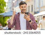 Small photo of Excited bearded man use mobile smartphone celebrating win good message news, success, lottery jackpot victory, giveaway online outdoor. Happy young guy tourist walking on urban city street. Lifestyles