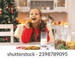 Happy joyful cute little girl in red sweater drinking milk and eating delicious cookie near Christmas tree in the kitchen for Christmas morning. Christmas time. Christmas holidays