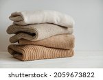 Bunch of knitted warm pastel color sweaters with different knitting patterns stacked in messy pile on white wooden table, white wall background. Fall winter season knitwear. Close up, copy space.