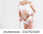 Small photo of Unrecognizable fit woman taking her body measurements with a centimeter measuring tape. Torso of a slim female with flat belly in white underwear. Copy space, isolated, white background, close up.