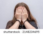 Small photo of A young girl handcuffed hides her face on a gray background, close-up. Juvenile delinquent in a black T-shirt, criminal liability of minors.