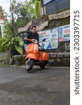 Small photo of Yogyakarta, Indonesia - Dec 31 2022: A man poses with his bright orange vespa strada aom motorcycle in a village parking space. Translate: mbayung green lombok stall benny, skilled in jogja