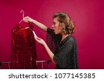 Shocked woman looking at price on sparkle red dress in boutique. Indoor portrait of amazed young female model holding hanger with expensive attire.