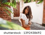 Woman touches her curly long hair. Tanned girl in bright summer outfit posing in resort town with tropical plants