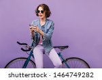 Inspired woman in casual attire looking at phone screen. Joyful white girl in dark sunglasses using smartphone during photoshoot with bicycle.