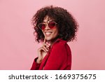 Charming girl in red jacket and bright glasses is smiling on pink background. Curly woman in stylish outfit posing on isolated backdrop