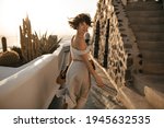 Attractive brunette woman in beige dress holds straw hat and runs along path in old beautiful city. Happy girl smiles and looks into camera.