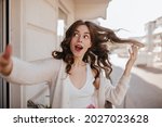 Small photo of Portrait shot of cute ukrainian lady playing with dark hair and touching camera. Dreaming of summer trips on the daylight veranda in genteel white clothes next to bouquet of flowers