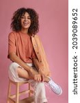 Small photo of stunning colorful lady of exotic appearance sits on high chair on light pink background. girl has bright snow-white smile, curly lush hair.