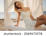 Young graceful lady sits on beach bed with white canopy on seashore. Pretty blonde gently tilts her head back, dressed in light summer dress. Elegant style, spring fashion trends.