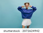 Happy young european lady in transparent glasses smiles with teeth at camera with place for text. Pretty woman with two blond buns on her head in blue sweatshirt and short white skirt.