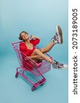 Small photo of Young caucasian woman in red dress rides inside shopping basket on blue background. Fair-haired model is having good time with nice mood. Relaxed freelance lifestyle, concept.