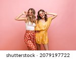 Small photo of Photo of two young caucasian women in bright clothes with polka dots standing on pink background. Blonde and brunette in sunglasses are smiling broadly with their teeth and showing sign of peace.