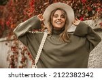 Attractive stylish woman holding hat on head, walking in park, dressed in warm suit. Pretty woman with light brown hair smiles broadly teeth. Autumn fashion accessories in street fashion style.