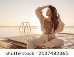 Smiling young caucasian girl, in light suit, trains on beach by sea. Woman with straight dark hair closed her eyes and turned head to side. Rest time concept.