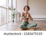Small photo of Pretty young brunette woman practicing yoga at home . Caucasian cute girl looking away and smiling wearing green sportswear sitting on the floor. Concept of leisure, relaxation, workout
