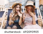 Small photo of Beautiful girls in glad rags celebrating an important event spending time outdoor. Portrait of funny female friends posing with surprised smile while drink champagne sitting on recliner.