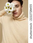 Small photo of Close-up photo.A beauteous young man with fair skin, with dark, short, beautiful hair combed back, in a beige turtleneck with a high collar stands on a dark beige background with a bouquet of daisies