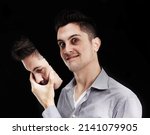 Small photo of Lurking insanity. Young man lifting a mask off his face and revealing a deranged expression.