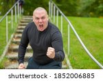 Small photo of Angry man threatening the camera with his fist as he screams abuse with a vehement expression on outdoors steps in a park