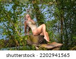 Small photo of Attractive blond woman wearing a gold gogo dress posing on a tree trunk at the edge of a lake