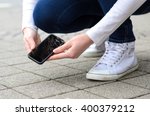 Close up view on kneeling person in jeans and shoes picking up broken phone on stone paved sidewalk outdoors