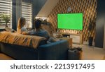 Small photo of Couple sitting on sofa in living room, talking, watching breaking news, movie or series on TV. Wife and husband resting and chilling at home in the evening together. Green screen. Chromakey.